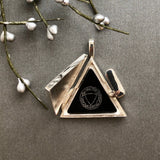 Triangular pendant with black center and emblem on a gray surface with willow branches.